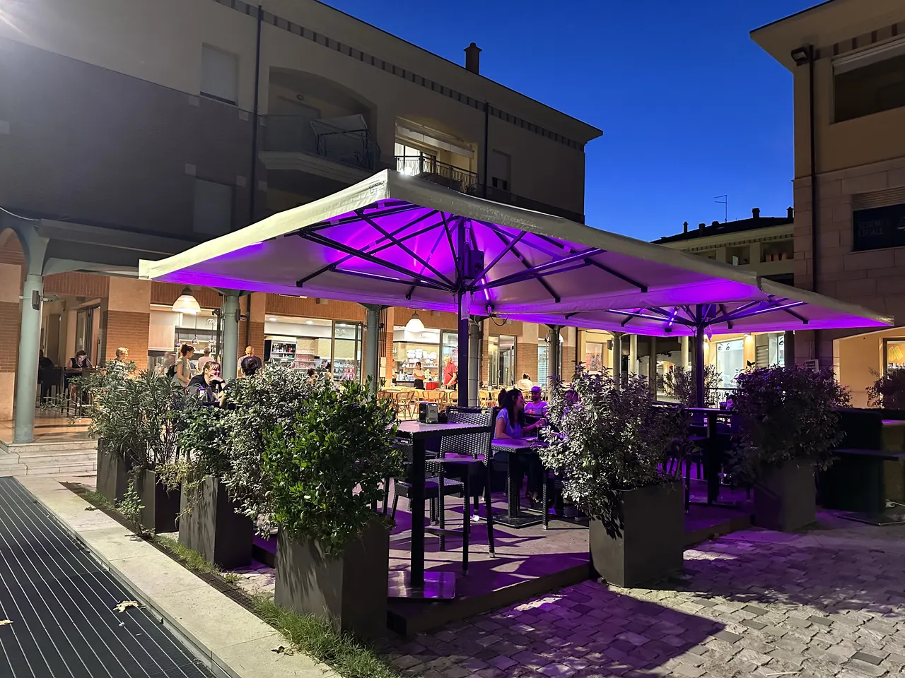 La piazza all'imbrunire: gli ombrelloni del Gabrè illuminati di viola, i primi tavoli dell'aperitivo